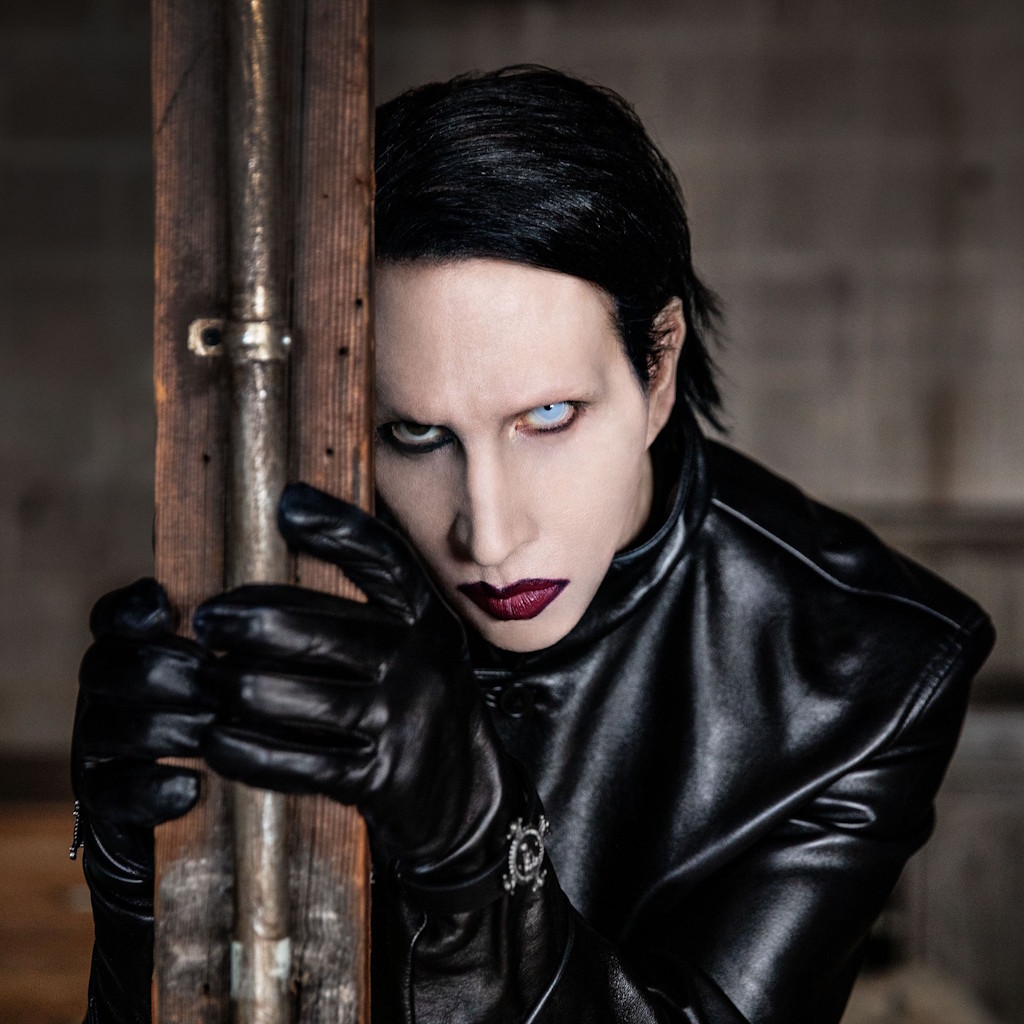 A portrait of Marilyn Manson. Manson is shown in a dramatic portrait, wearing a glossy black leather jacket, matching leather gloves, and heavy makeup. His left eye is light, electric blue, and his right eye is significantly darker. He leans against a vertical wooden girder with a powerline running up it, staring intently into the camera, showcasing his iconic bold lipstick and slicked-back black hair. The background is dimly lit and out of focus, highlighting his gothic attire and enigmatic presence.