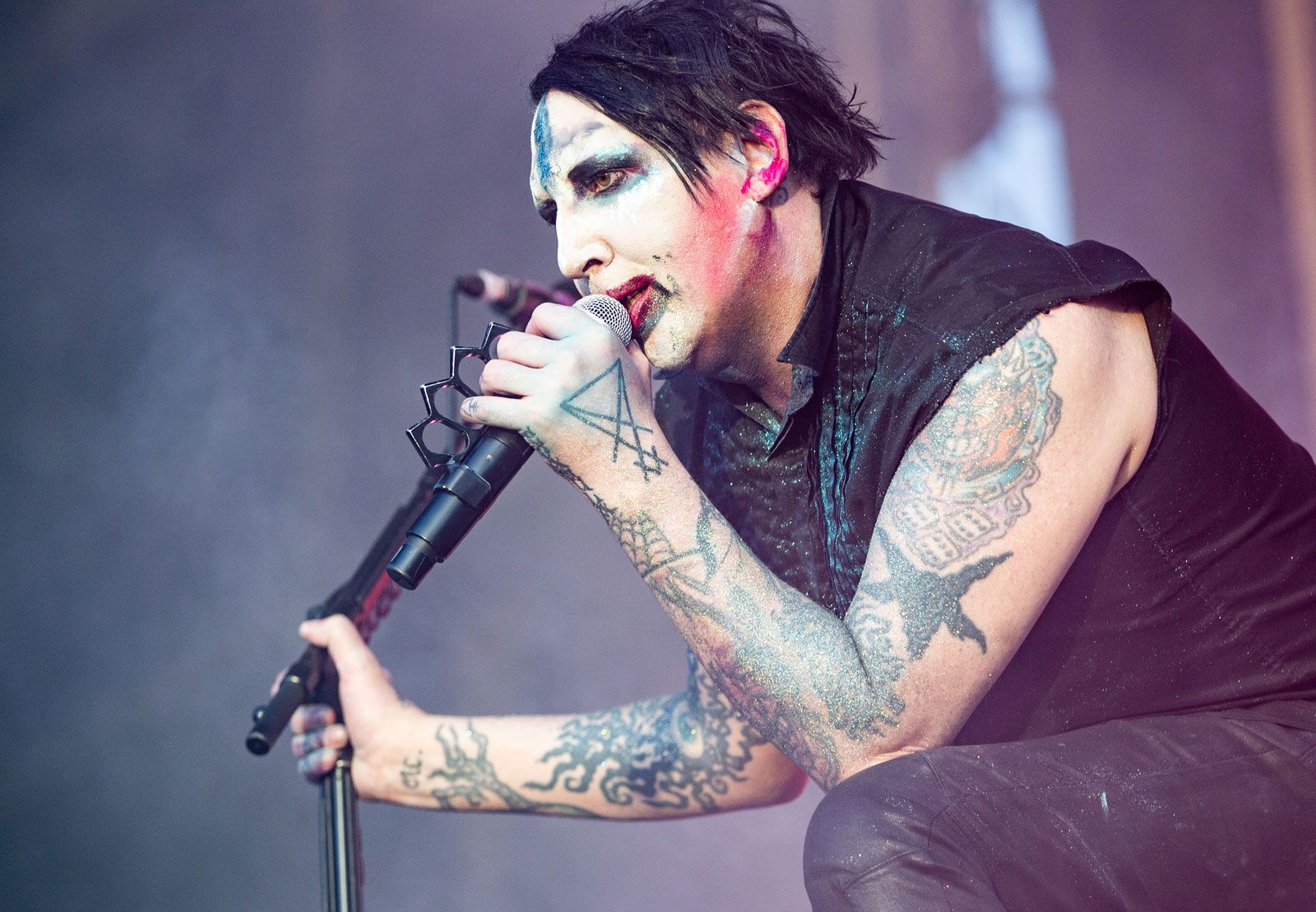 The image captures a raw and electrifying moment of Marilyn Manson in performance, showcasing his signature theatrical style. Manson, with his iconic painted face and striking makeup, grips the microphone with intensity and poise. His tattoos are vividly displayed, adding to his distinctive look. The lighting casts a mystical glow around him, highlighting his captivating presence on stage. This portrait reflects the essence of Manson's enigmatic artistry and his unparalleled ability to captivate audiences worldwide with his provocative performances.