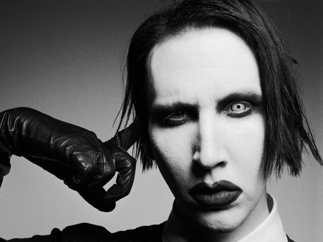 In this striking black-and-white portrait, Marilyn Manson exudes an aura of provocative elegance and intensity. His face, stark and pale, is accentuated by bold, dark lipstick and sharply defined eye makeup that underscores his penetrating stare. Manson's gloved hand is poised against his temple, adding a dramatic touch to his enigmatic expression. The composition is minimalistic yet potent, capturing the essence of Manson's avant-garde persona and his impactful presence in the realm of music and art. This image epitomizes his unique style and the fearless self-expression that defines his artistic legacy.